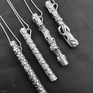 May include: Five silver necklaces with different pendant designs. Each pendant is a long, thin cylinder with intricate carvings. The pendants feature skulls, flowers, and a tree-like design.