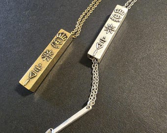 May include: Two pendant necklaces. One is gold-toned, the other silver-toned. Each has a rectangular pendant with engraved symbols and hangs from a chain. The silver necklace has a separate, bar-shaped pendant. The pendants are likely jewelry.