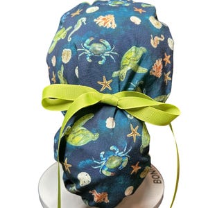 May include: A dark blue scrub cap featuring a sea life pattern with crabs, turtles, starfish, and seahorses. The cap is tied with a bright green ribbon. The design includes coral and seashells. The cap is on a white stand.