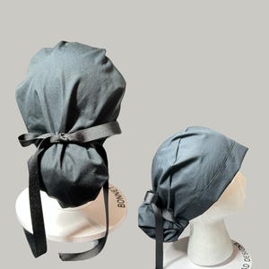 May include: Two dark gray bonnets, one displayed upright and the other on a mannequin head. Both bonnets have a ribbon tie. The bonnets are on white circular stands with the text "BONNET HEAD DESIGNS".