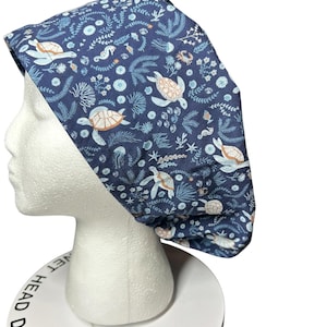 May include: A navy blue scrub cap featuring a sea life pattern. The design includes sea turtles, seahorses, and various aquatic plants in shades of blue, white, and tan. The cap is displayed on a white mannequin head.