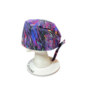 May include: A patterned surgical scrub cap featuring a swirling galaxy design in purple, blue, pink, and black. The cap has matching fabric ties at the back. It is displayed on a white mannequin head.