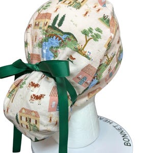 May include: A white bonnet with a repeating pattern of houses, trees, and cows. The bonnet is tied with a green ribbon. The bonnet is displayed on a white mannequin head.