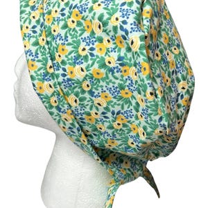 May include: A floral scrub cap featuring a pattern of yellow roses, blue accents, and green leaves on a light green background. The cap is tied at the back with two long ties. The cap is designed to be worn on the head.