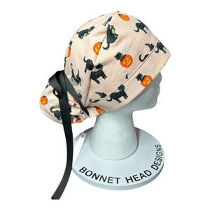 May include: A light peach-colored scrub cap featuring a Halloween-themed pattern. The design includes black cats, jack-o'-lanterns, and witch hats. The cap has a black ribbon tie and is displayed on a white mannequin head.