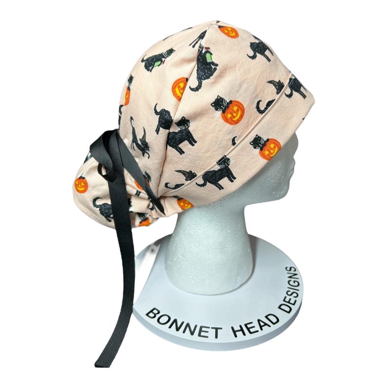 May include: A light peach-colored scrub cap featuring a Halloween-themed pattern. The design includes black cats, jack-o'-lanterns, and witch hats. The cap has a black ribbon tie and is displayed on a white mannequin head.