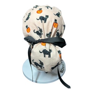 May include: A light pink scrub cap featuring a black cat and jack-o'-lantern pattern. The cap has a black ribbon tie and is designed for medical professionals. The Halloween-themed design adds a festive touch to work attire.