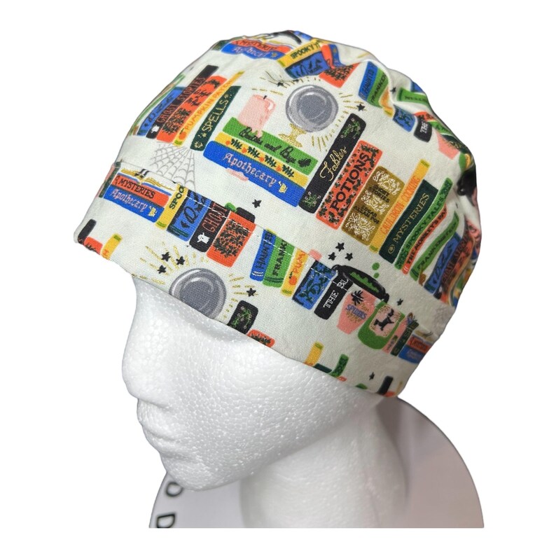 K&ouml;nnte beinhalten: Eine wei&szlig;e OP-Haube mit einem farbenfrohen Aufdruck von gestapelten B&uuml;chern, Tr&auml;nken und Apothekenetiketten. Die Haube zeigt Texte wie "Zauberspr&uuml;che", "Tr&auml;nke" und "Geheimnisse". Das Design umfasst Sterne und kreisf&ouml;rmige Akzente.