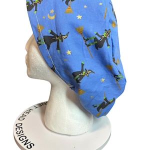 May include: A blue scrub cap featuring a repeating pattern of witches flying on broomsticks, accented with yellow stars and crescent moons. The fabric displays black-clad witches with green skin and striped stockings against a light blue background.