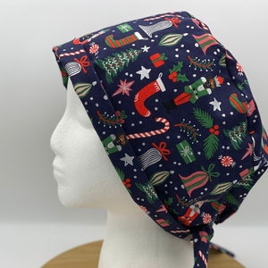 May include: A navy blue surgical scrub cap with a festive Christmas pattern. The fabric is covered with illustrations of Christmas trees, stockings, nutcrackers, and candy canes in red, green, and white. The cap is tied at the back.
