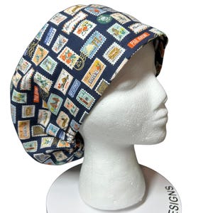May include: A navy blue bonnet with a repeating pattern of colorful postage stamps. The bonnet is displayed on a white mannequin head. Text on the bonnet includes "PAR AVION", "FIRST CLASS POSTAGE", and "SPECIAL DELIVERY". The base reads "BONNET HEAD DESIGNS".
