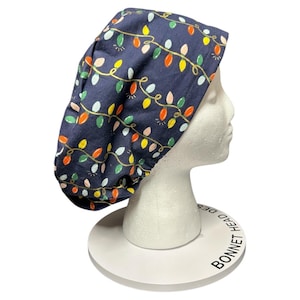 May include: A navy blue scrub cap featuring a repeating pattern of colorful Christmas lights. The lights are in shades of red, yellow, green, and pink, connected by a gold-colored wire. The cap is displayed on a white mannequin head.