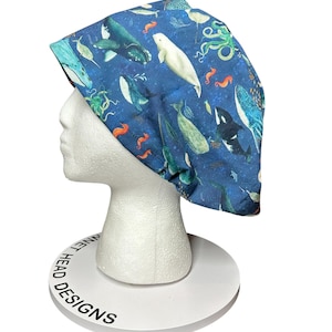May include: A blue scrub cap featuring an all-over print of various marine animals, including whales, octopuses, and seahorses. The cap is displayed on a white mannequin head. The text "BONNET HEAD DESIGNS" is printed on the base.