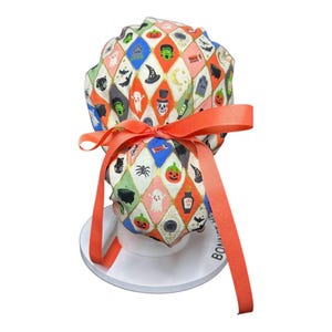 May include: A colorful Halloween-themed scrub cap with a diamond pattern featuring pumpkins, ghosts, and bats. The cap is tied with a bright orange ribbon. The cap is on a white stand.