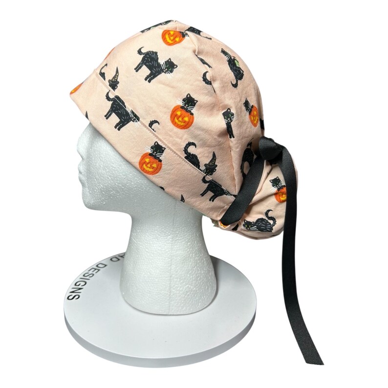 May include: A light pink Halloween scrub cap featuring a repeating pattern of black cats, jack-o'-lanterns, and witch hats. The cap has a black ribbon tie and is displayed on a white mannequin head. The cap is designed for medical professionals.