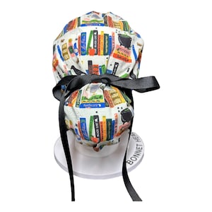 K&ouml;nnte beinhalten: Eine wei&szlig;e OP-Haube mit schwarzer Schleife und Bindeb&auml;ndern. Die Haube ist mit bunten B&uuml;chern, Zaubertr&auml;nken und den W&ouml;rtern "Spells", "Potions" und "The Black Cat" bedruckt.
