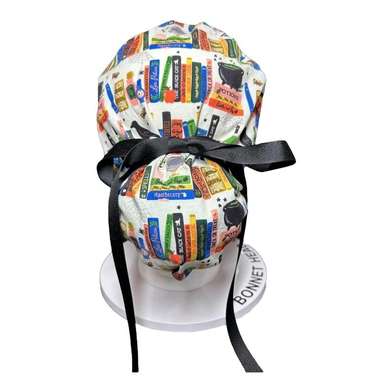 K&ouml;nnte beinhalten: Eine wei&szlig;e OP-Haube mit schwarzer Schleife und Bindeb&auml;ndern. Die Haube ist mit bunten B&uuml;chern, Zaubertr&auml;nken und den W&ouml;rtern "Spells", "Potions" und "The Black Cat" bedruckt.