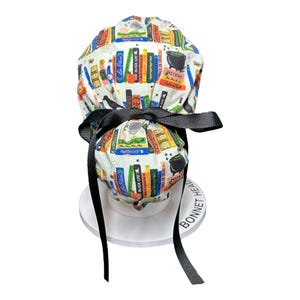 K&ouml;nnte beinhalten: Eine wei&szlig;e OP-Haube im Bonnet-Stil mit einer schwarzen Schleife. Die Haube ist mit bunten Buchr&uuml;cken, Zaubertr&auml;nken und dem Text "Potions", "Spells" und "Black Cat" gemustert. Die Haube ist mit einem schwarzen Band gebunden.