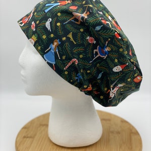May include: A dark green scrub cap featuring a festive Nutcracker-themed pattern. The fabric is covered with illustrations of ballerinas, toy soldiers, candy canes, and other holiday elements. The cap is designed to fit comfortably and securely.
