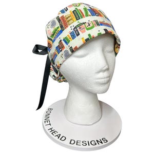 K&ouml;nnte beinhalten: Eine wei&szlig;e OP-Haube mit einem bunten Muster aus B&uuml;chern und Kaffeetassen. Die Haube hat eine schwarze Schleife und wird auf einem wei&szlig;en Schaufensterkopf pr&auml;sentiert. Auf dem Sockel des Schaufensterkopfes steht "Bonnet Head Designs".