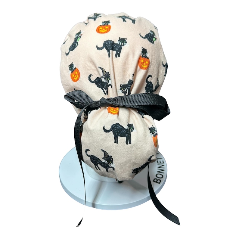 May include: A light pink Halloween scrub cap featuring a repeating pattern of black cats and orange jack-o'-lanterns. The cap is tied with a black ribbon and is displayed on a white stand. The cap is designed for medical professionals.