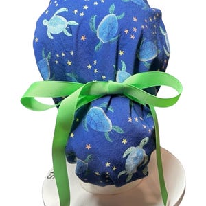 May include: A blue scrub cap featuring a pattern of light blue sea turtles and yellow stars. The cap is tied with a bright green ribbon. The cap is designed to be worn in a medical or veterinary setting.