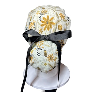 Peut inclure: Une coiffe de bloc opératoire à motifs floraux blancs et jaunes, avec un nœud noir. Le motif de la coiffe comprend des abeilles, des fleurs et des feuilles. Un nœud noir est noué à l'arrière, avec de longs rubans noirs.