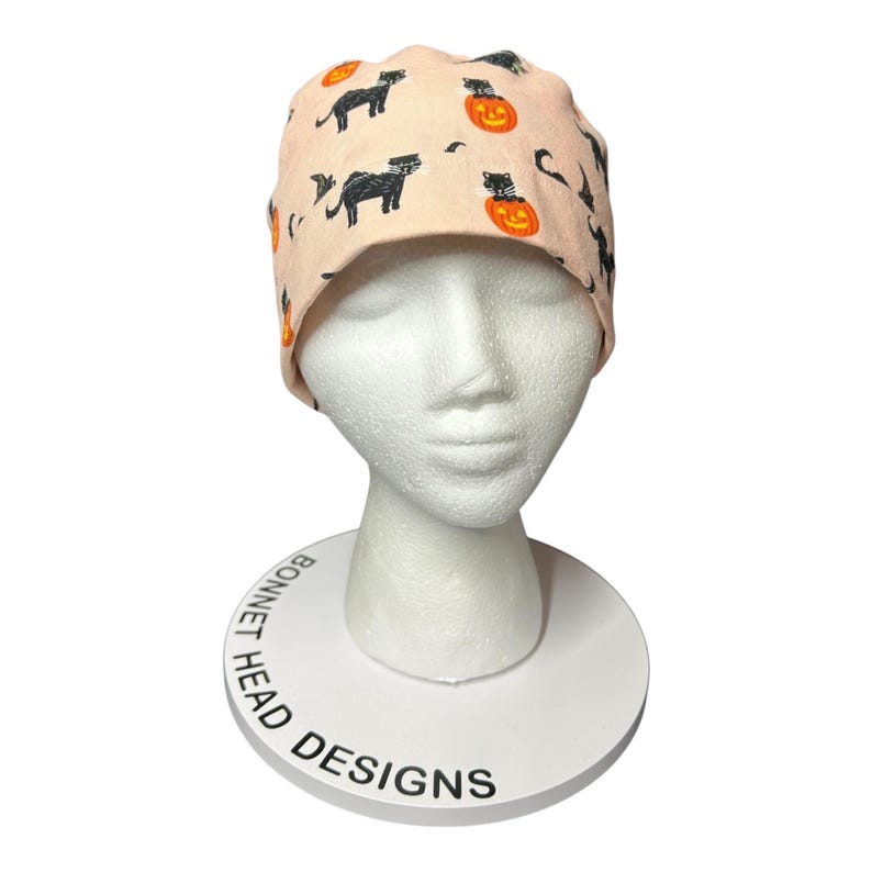 May include: A light peach-colored surgical scrub cap with a Halloween-themed pattern. The pattern includes black cats, orange jack-o'-lanterns, and bats. The cap is displayed on a white mannequin head.