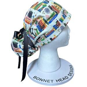 K&ouml;nnte beinhalten: Eine wei&szlig;e OP-Haube mit einem bunten Aufdruck von Zaubertr&auml;nken, B&uuml;chern und Apothekenetiketten. Die Haube hat eine schwarze Schleife und wird auf einem wei&szlig;en Schaufensterkopf pr&auml;sentiert. Der Text "POTIONS" und "BONNET HEAD DESIGNS" sind sichtbar.