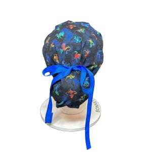 May include: A dark blue scrub cap with a pattern of colorful dragons. The cap is tied with a bright blue ribbon. The cap is on a white stand.