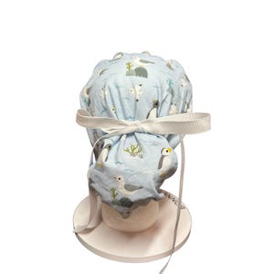 May include: A light blue scrub cap featuring a pattern of penguins, seagulls, and aquatic plants. The cap is tied with a white ribbon bow. The cap is designed for medical professionals and is made of a soft, breathable fabric.