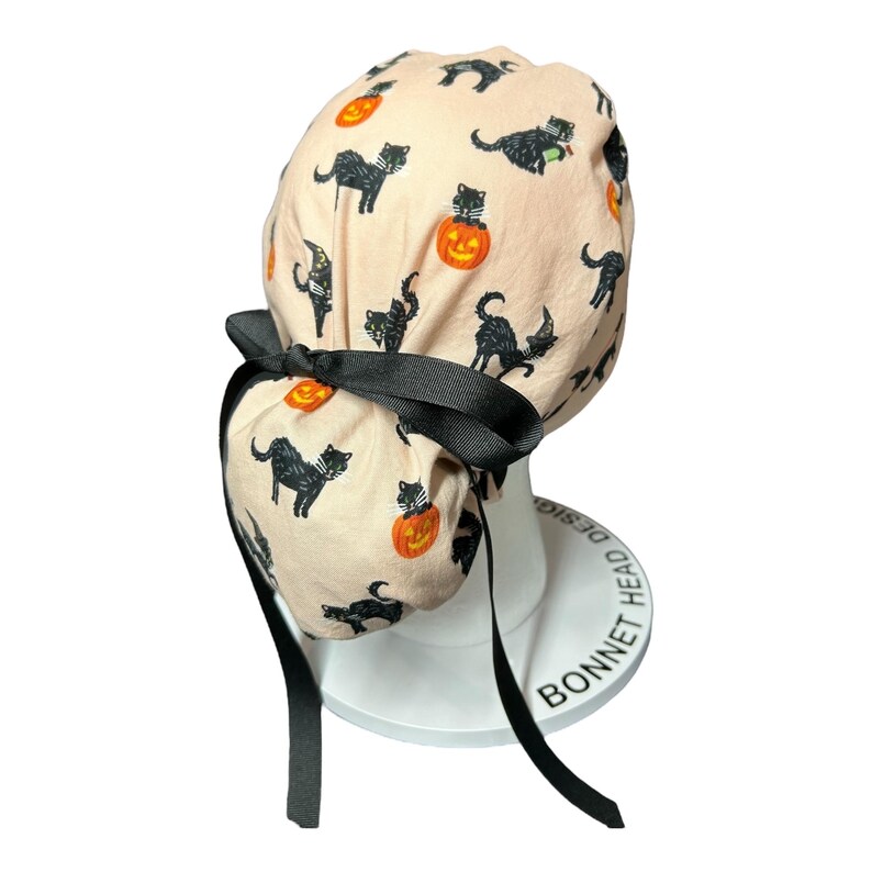 May include: A light peach-colored bonnet featuring a Halloween-themed pattern of black cats and jack-o'-lanterns. The bonnet is tied with a black bow and has long black ribbons. The bonnet is designed to be worn on the head.