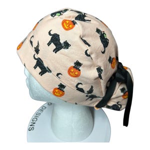 May include: A light peach-colored scrub cap featuring a Halloween-themed pattern. The design includes black cats, orange jack-o'-lanterns, and witch hats. The cap is tied with a black ribbon. The words "P Designs" are visible on the base.