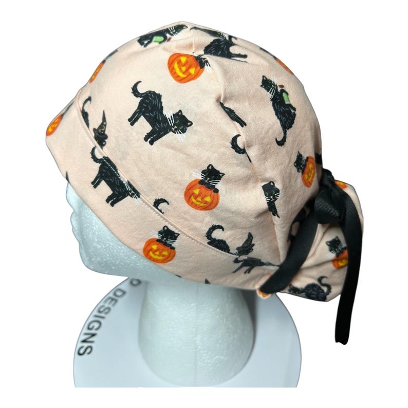 May include: A light peach-colored scrub cap featuring a Halloween-themed pattern. The design includes black cats, orange jack-o'-lanterns, and witch hats. The cap is tied with a black ribbon. The words "P Designs" are visible on the base.