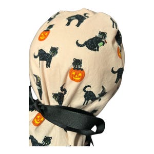 May include: A light beige Halloween scrub cap featuring a repeating pattern of black cats and orange jack-o'-lanterns. The cap has a black ribbon tie. The cats are in various poses, some with green eyes.