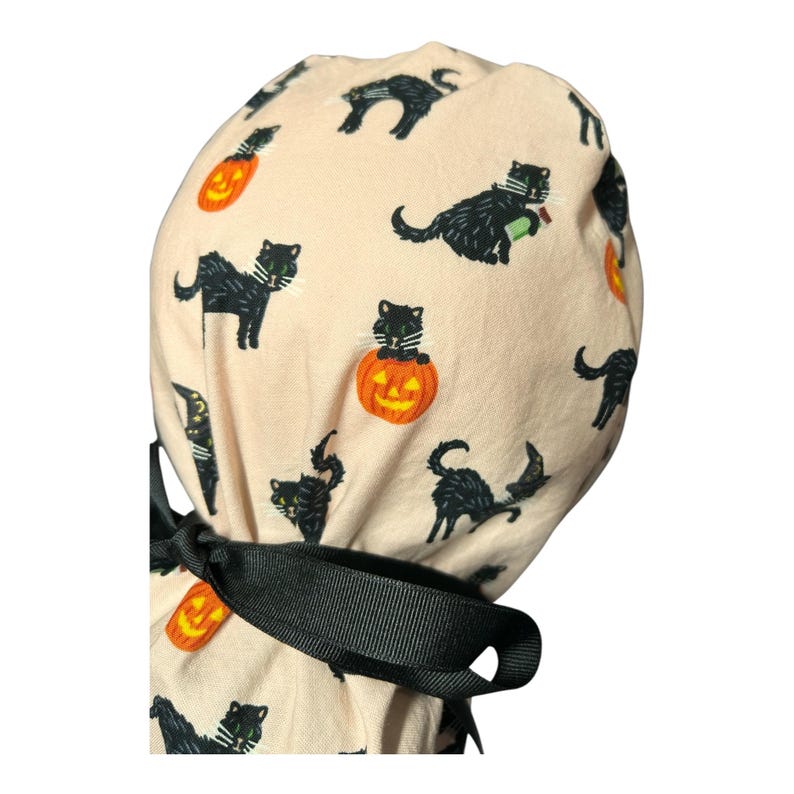 May include: A light beige Halloween scrub cap featuring a repeating pattern of black cats and orange jack-o'-lanterns. The cap has a black ribbon tie. The cats are in various poses, some with green eyes.