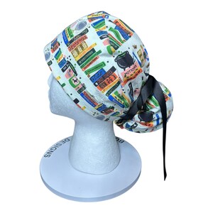 K&ouml;nnte beinhalten: Eine wei&szlig;e OP-Haube mit einem farbenfrohen Aufdruck von gestapelten B&uuml;chern, Zaubertr&auml;nken und Kesseln. Die Haube hat eine schwarze Schleife und wird auf einem wei&szlig;en Schaufensterkopf pr&auml;sentiert. Der Text auf den B&uuml;chern lautet "Potions", "Mysteries" und "Frankenstein".