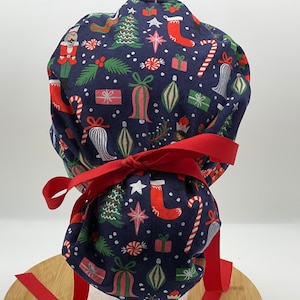 May include: A navy blue scrub cap with a festive Christmas pattern. The fabric features red, green, and white holiday icons like Santa, ornaments, candy canes, and gifts. A red ribbon is tied around the cap.