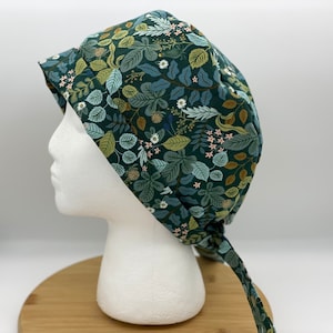 May include: A dark green scrub cap with a botanical pattern featuring leaves, berries, and small flowers in shades of green, blue, and pink. The cap is tied at the back with long fabric ties.