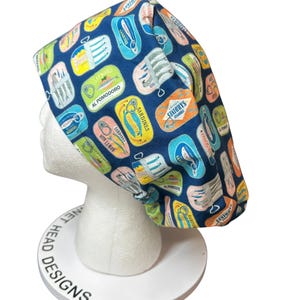 May include: A scrub cap featuring a dark blue background with a repeating pattern of colorful sardine tin illustrations. The tins are depicted in various shades of yellow, orange, light blue, and white, each labeled with text such as Sardines, Al Pomodoro, and With Lemon.