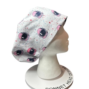 May include: A white bonnet with a repeating pattern of pink astronaut helmets and red and blue stars. The bonnet is displayed on a white mannequin head and a white stand with the text "BONNET HEAD DESIGNS".