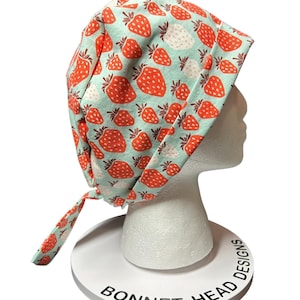 May include: A light blue bonnet with a repeating pattern of red and white strawberries. The bonnet is tied at the back and is displayed on a white mannequin head. The text "BONNET HEAD DESIGNS" is printed on the base.
