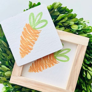 May include: A wooden framed sign with a hand-painted orange carrot with green leaves. The sign is sitting on a bed of green boxwood.