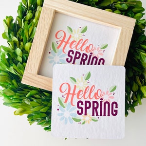 May include: Two wooden framed signs with the text "Hello Spring" in colorful lettering and floral designs. The signs are on a green background.