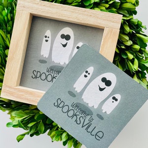 May include: A gray welcome sign with the text "Welcome to Spooksville" and a group of three white ghosts with black eyes and smiles. The sign is framed in a light wood frame.