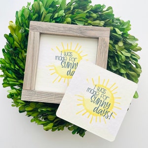May include: A framed sign and a square coaster with a yellow sun design and the text "I was made for sunny days!" The sign is in a gray wooden frame. The items are set on a bed of green leaves.
