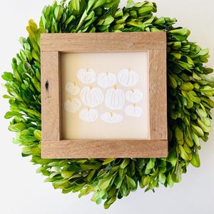 Puede incluir: Una impresi&oacute;n de arte enmarcada con un fondo blanco y un grupo de calabazas blancas con tallos naranjas. El marco es de madera y tiene un acabado natural.