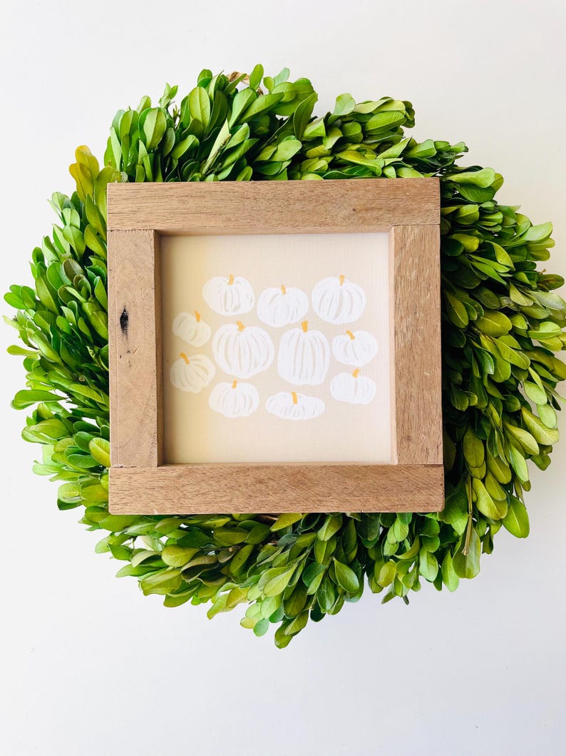 Puede incluir: Una impresi&oacute;n de arte enmarcada con un fondo blanco y un grupo de calabazas blancas con tallos naranjas. El marco es de madera y tiene un acabado natural.