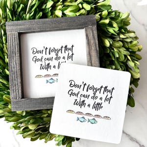 May include: Two white signs with the text "Don't forget that God can do a lot With a little" in black script. The signs have illustrations of bread and fish. One sign is framed in gray wood.