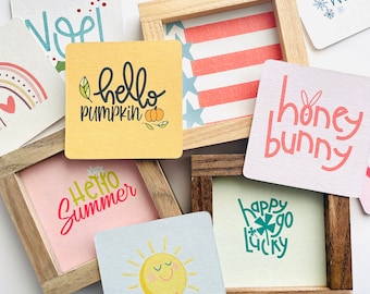 May include: A collection of colorful square cards with various greetings and designs. The cards feature phrases like "hello pumpkin", "hello summer", "honey bunny", "happy go lucky", "you are my sunshine", "back to school", and "hello winter". Some cards are framed in wood.